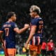 Two soccer players in navy and orange uniforms shake hands on the field as a match continues, with a camera to the right and a blurred crowd in the stands behind them.