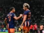 Two soccer players in navy and orange uniforms shake hands on the field as a match continues, with a camera to the right and a blurred crowd in the stands behind them.