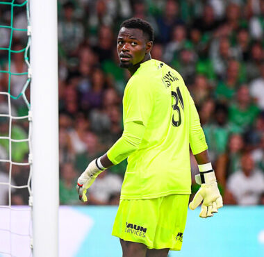 Soccer goalkeeper in bright neon-green kit stands near the goal, looking back over his shoulder as the crowd watches.