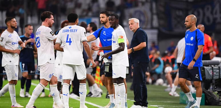 Soccer players in white uniforms shake hands and chat on the sideline as a coach watches on a green field.