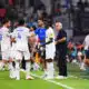 Soccer players in white uniforms shake hands and chat on the sideline as a coach watches on a green field.