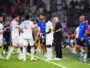 Soccer players in white uniforms shake hands and chat on the sideline as a coach watches on a green field.
