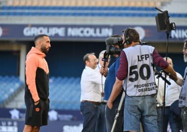 Athlete in a peach hoodie being interviewed by a TV crew with cameras at a stadium backdrop.