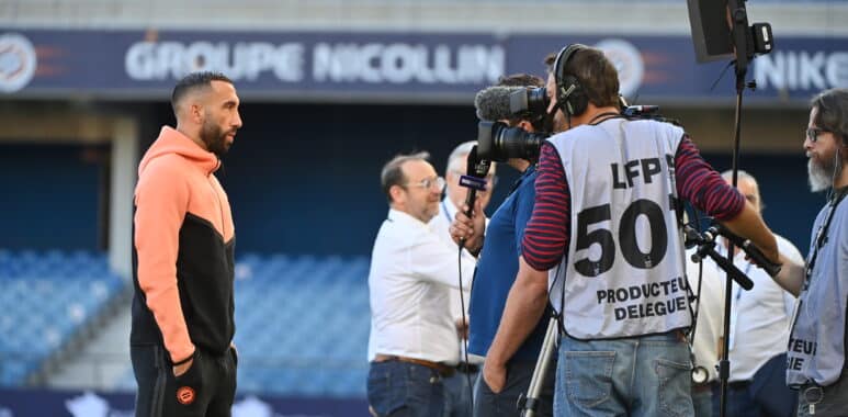 Athlete in a peach hoodie being interviewed by a TV crew with cameras at a stadium backdrop.