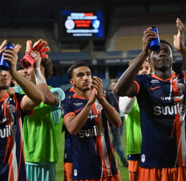 Soccer players in blue and orange striped uniforms celebrate, raising blue water bottles on the field.