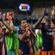 Soccer players in blue and orange striped uniforms celebrate, raising blue water bottles on the field.