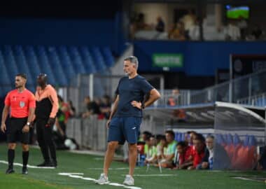 Soccer coach in blue kit stands on the sideline with hands on hips as players and spectators watch from the background.