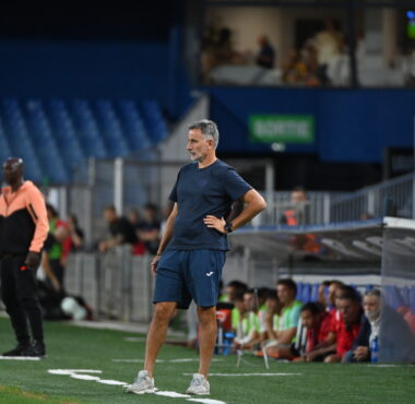 Soccer coach in blue kit stands on the sideline with hands on hips as players and spectators watch from the background.