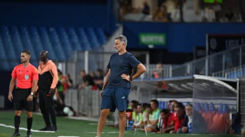 Soccer coach in blue kit stands on the sideline with hands on hips as players and spectators watch from the background.