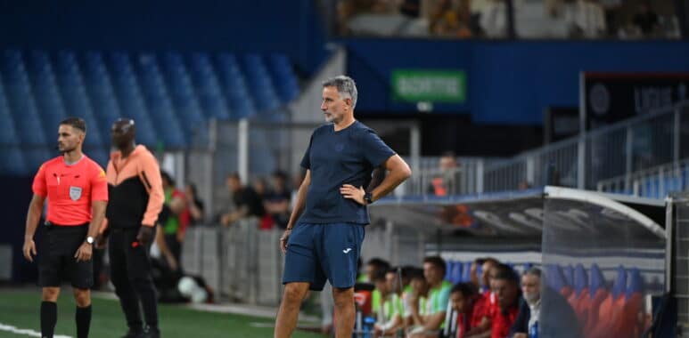 Soccer coach in blue kit stands on the sideline with hands on hips as players and spectators watch from the background.