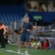 Soccer coach in blue kit stands on the sideline with hands on hips as players and spectators watch from the background.