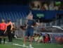 Soccer coach in blue kit stands on the sideline with hands on hips as players and spectators watch from the background.