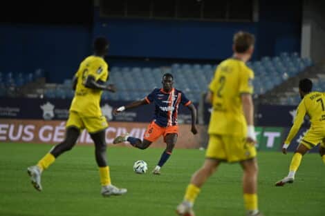 Soccer player in dark blue and orange kit dribbles the ball while opponents in yellow close in on him on a green field.