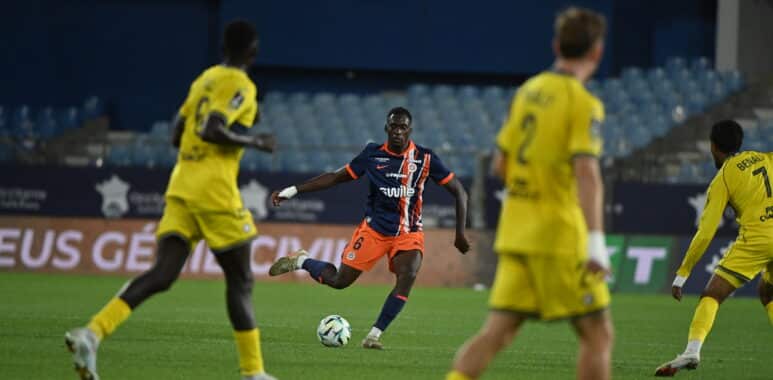 Soccer player in dark blue and orange kit dribbles the ball while opponents in yellow close in on him on a green field.