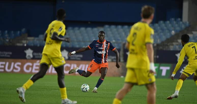 Soccer player in dark blue and orange kit dribbles the ball while opponents in yellow close in on him on a green field.