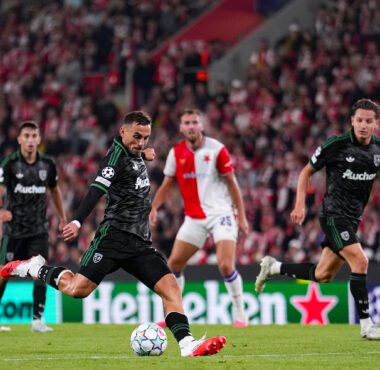Soccer players chase a ball on a crowded, red-clad stadium; central player in black kicks forward while teammates run nearby.