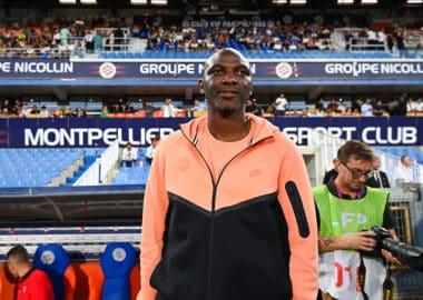 Athlete standing on a stadium sideline wearing a peach and black zip-up jacket, with fans in the stands behind him.