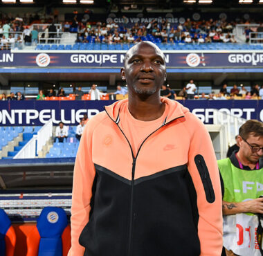 Athlete standing on a stadium sideline wearing a peach and black zip-up jacket, with fans in the stands behind him.