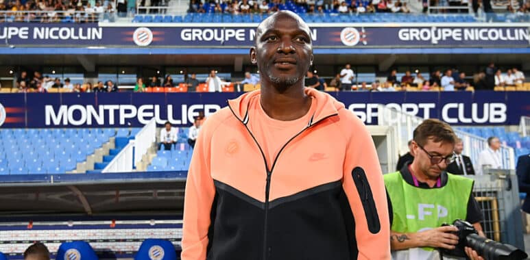 Athlete standing on a stadium sideline wearing a peach and black zip-up jacket, with fans in the stands behind him.