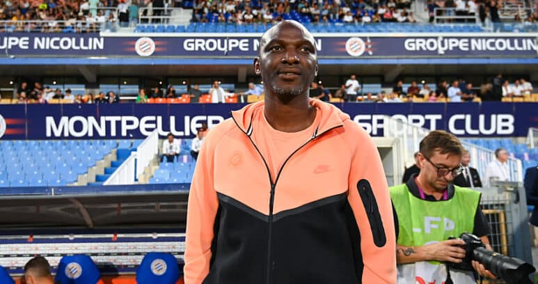 Athlete standing on a stadium sideline wearing a peach and black zip-up jacket, with fans in the stands behind him.
