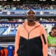 Athlete standing on a stadium sideline wearing a peach and black zip-up jacket, with fans in the stands behind him.