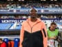 Athlete standing on a stadium sideline wearing a peach and black zip-up jacket, with fans in the stands behind him.