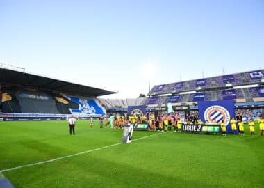 Soccer players line up on the green pitch before a match, with banners and a large stadium crowd in the background.