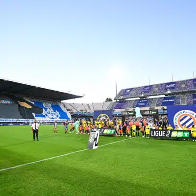 Soccer players line up on the green pitch before a match, with banners and a large stadium crowd in the background.