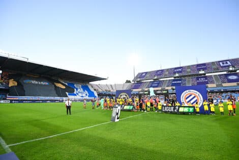 Soccer players line up on the green pitch before a match, with banners and a large stadium crowd in the background.