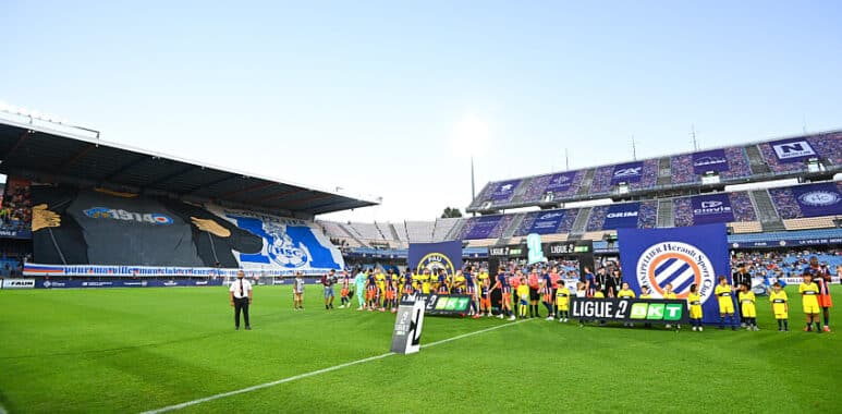 Soccer players line up on the green pitch before a match, with banners and a large stadium crowd in the background.