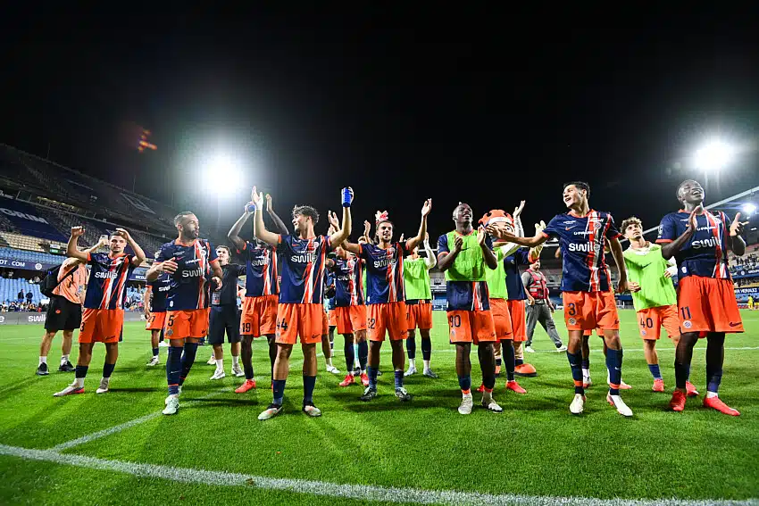 Soccer team in navy and orange uniforms applauds on a well-lit night field, arms raised in celebration.