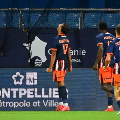 Three soccer players in blue and orange kits walk along the sideline, backs to the camera; jersey numbers 17 and 23 visible, with advertising boards in the background reading Montpellier M&eacute;tropole et Ville.