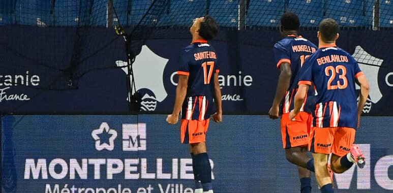 Three soccer players in blue and orange kits walk along the sideline, backs to the camera; jersey numbers 17 and 23 visible, with advertising boards in the background reading Montpellier M&eacute;tropole et Ville.
