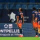 Three soccer players in blue and orange kits walk along the sideline, backs to the camera; jersey numbers 17 and 23 visible, with advertising boards in the background reading Montpellier M&eacute;tropole et Ville.