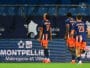 Three soccer players in blue and orange kits walk along the sideline, backs to the camera; jersey numbers 17 and 23 visible, with advertising boards in the background reading Montpellier M&eacute;tropole et Ville.