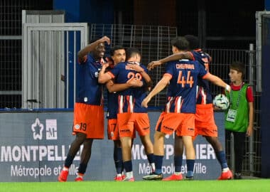 Chennaiyin FC players celebrate with a group hug on the field in blue and orange uniforms near advertising boards.