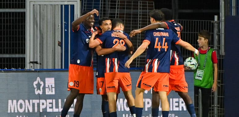 Chennaiyin FC players celebrate with a group hug on the field in blue and orange uniforms near advertising boards.