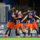 Chennaiyin FC players celebrate with a group hug on the field in blue and orange uniforms near advertising boards.