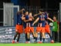 Chennaiyin FC players celebrate with a group hug on the field in blue and orange uniforms near advertising boards.