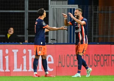 Two soccer players in navy and orange kits celebrate a goal with a high-five on the field, a spectator visible in the background near advertising boards.