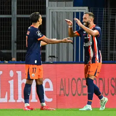 Two soccer players in navy and orange kits celebrate a goal with a high-five on the field, a spectator visible in the background near advertising boards.
