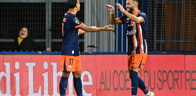 Two soccer players in navy and orange kits celebrate a goal with a high-five on the field, a spectator visible in the background near advertising boards.
