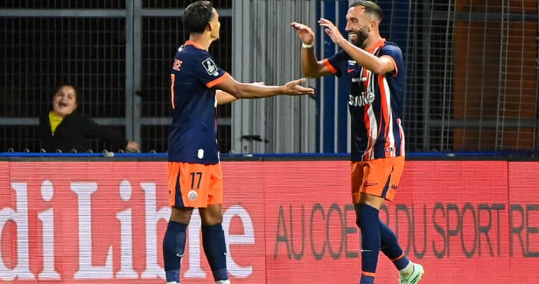 Two soccer players in navy and orange kits celebrate a goal with a high-five on the field, a spectator visible in the background near advertising boards.