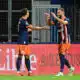 Two soccer players in navy and orange kits celebrate a goal with a high-five on the field, a spectator visible in the background near advertising boards.