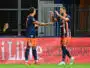 Two soccer players in navy and orange kits celebrate a goal with a high-five on the field, a spectator visible in the background near advertising boards.