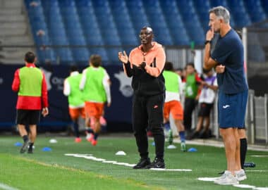 Coach in orange and black tracksuit gestures while speaking to players on a soccer field during training.