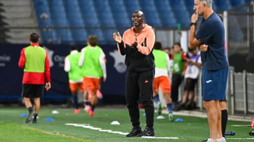 Coach in orange and black tracksuit gestures while speaking to players on a soccer field during training.