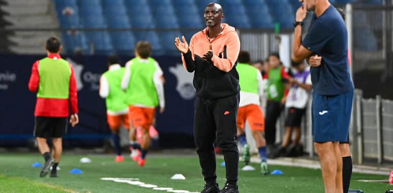 Coach in orange and black tracksuit gestures while speaking to players on a soccer field during training.