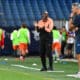 Coach in orange and black tracksuit gestures while speaking to players on a soccer field during training.