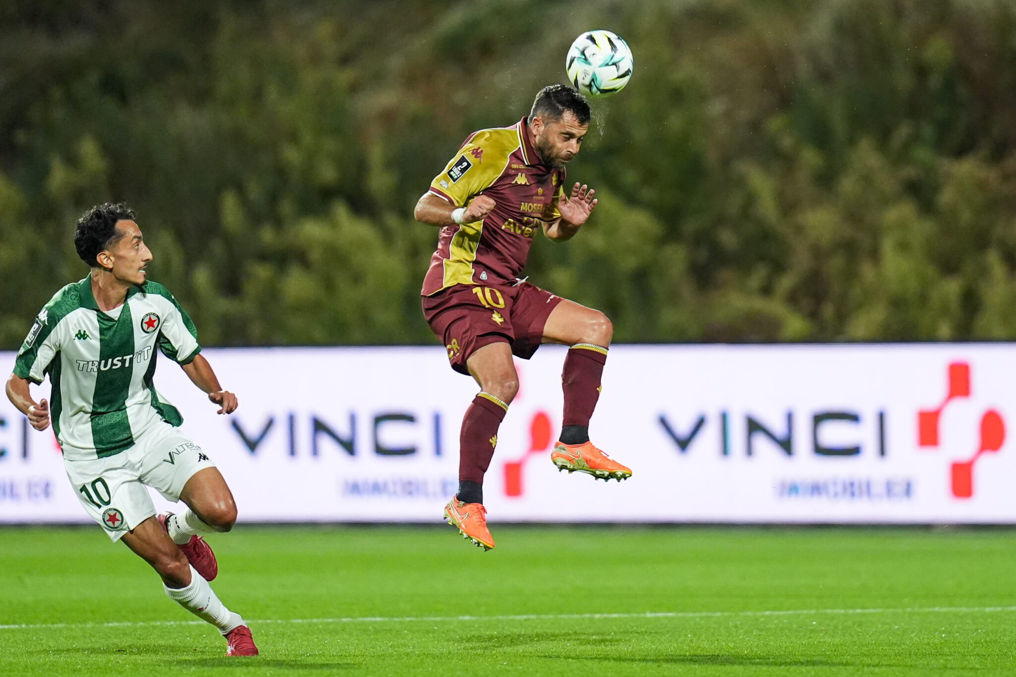 Téji Savanier Red Star FC v FC Metz - Ligue 2 BKT 20262027 | AllezPaillade.com Soccer players contesting a ball mid-air; maroon-clad striker jumps while a green-white opponent pursues on a grass field.